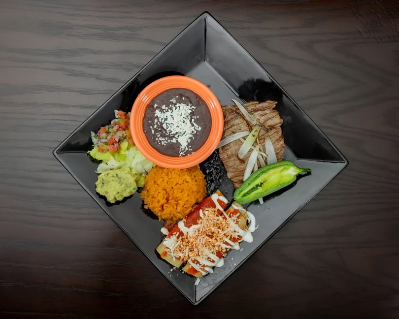 A black square plate holds a Mexican combo meal with steak, enchiladas, rice, beans, guacamole, and salsa.