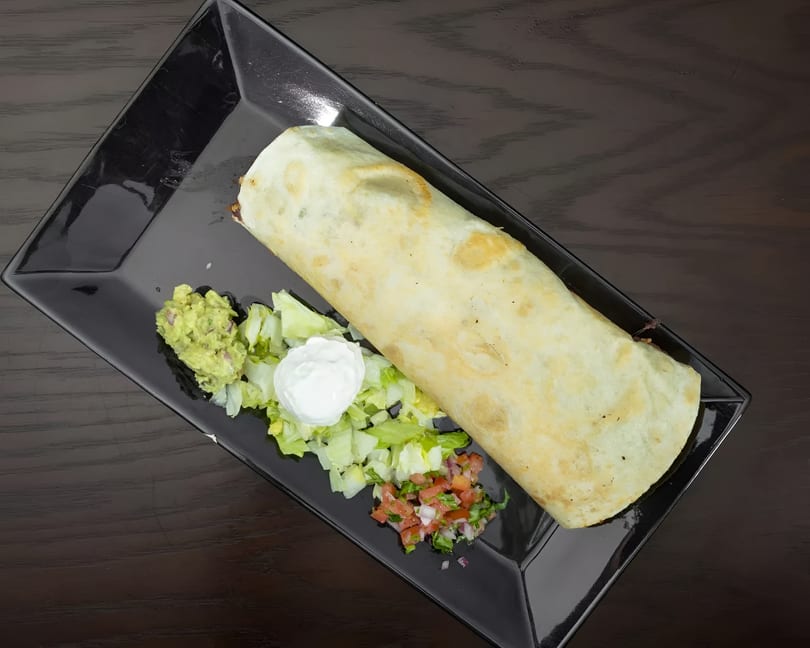 Large burrito on a black plate with sides of guacamole, shredded lettuce, sour cream, and pico de gallo.