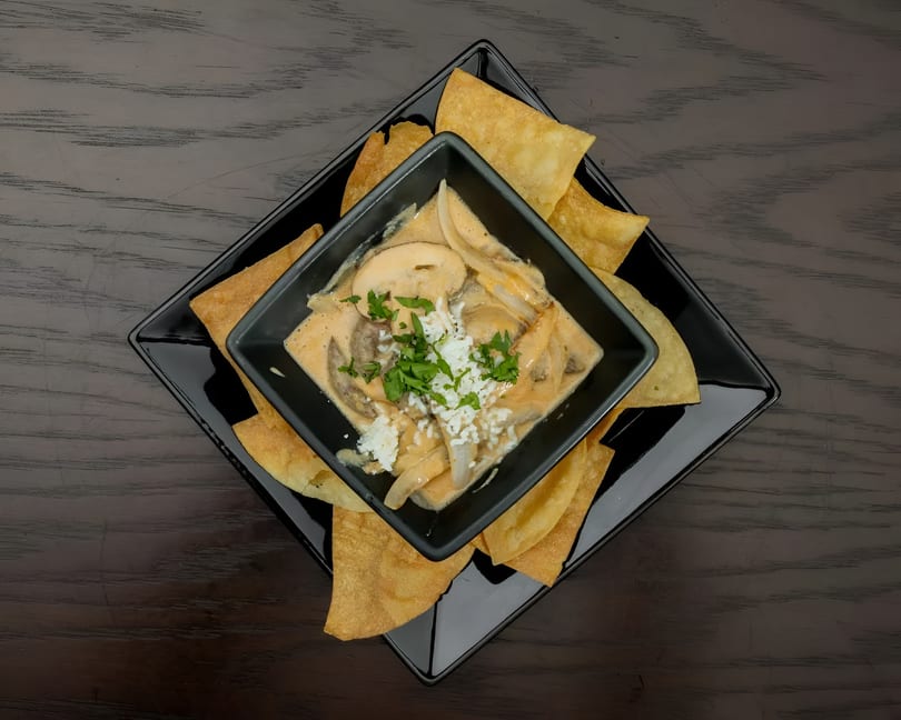 Mushroom and onion dip with crumbled cheese and parsley, served with tortilla chips on a black plate.