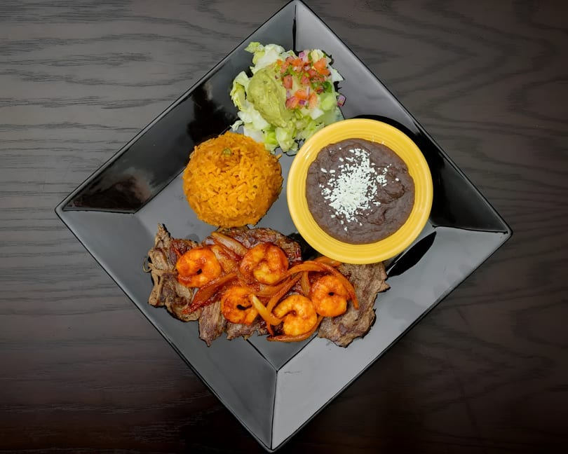 Grilled steak and shrimp with rice, refried beans, guacamole, and pico de gallo on a black plate.
