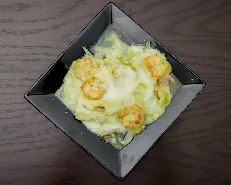 Shrimp dish with melted white cheese and green sauce on a black square plate.