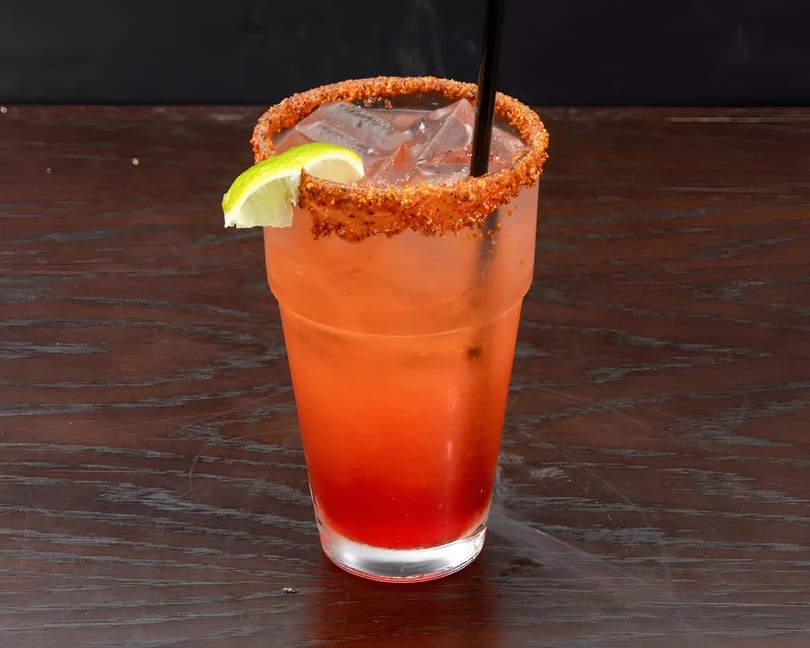 A red cocktail with a chili-lime rim, ice, a lime wedge, and a black straw on a wooden table.