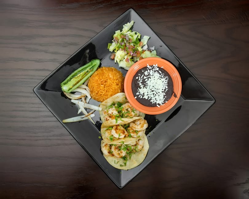 Three shrimp tacos with rice, black beans, salad, grilled jalapeño, and onions on a black plate.