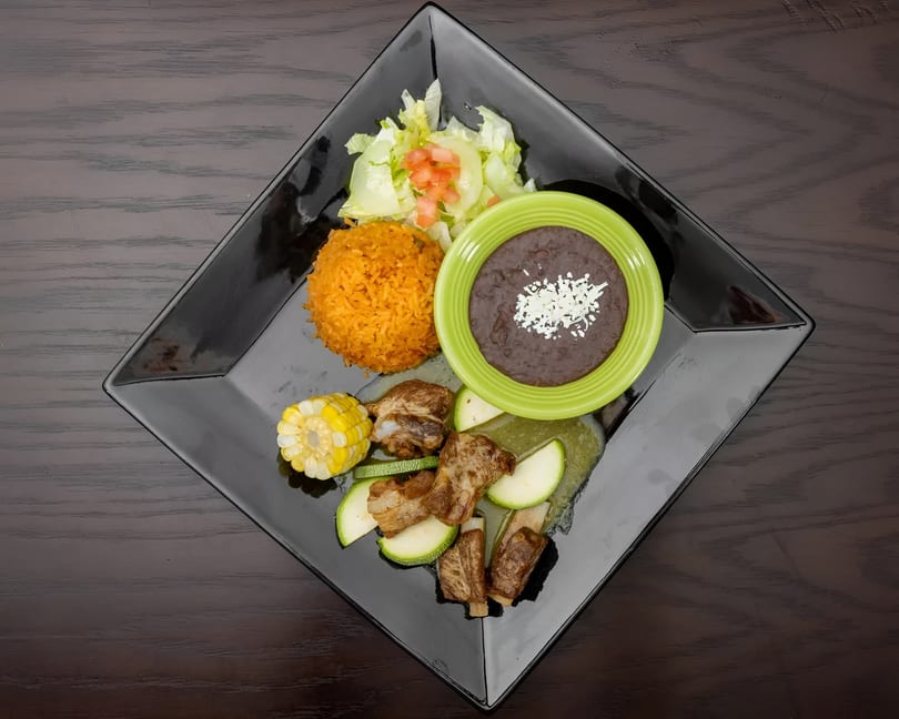 A black square plate with ribs, zucchini, corn, rice, salad, and a bowl of black beans.