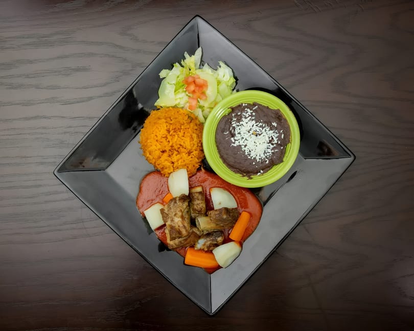 Mexican dish with ribs in red sauce, rice, refried beans, and a side salad on a black plate.