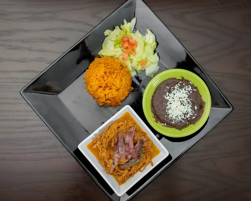 A black square plate holds a Mexican meal with rice, salad, refried beans, and cochinita pibil.