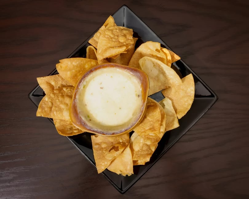 A black square plate holds a bowl of creamy queso dip surrounded by crispy tortilla chips.