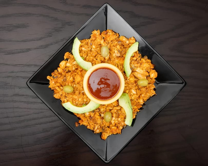 A black square plate holds a mound of orange-brown food, garnished with avocado slices, green olives, and a small bowl of red sauce.