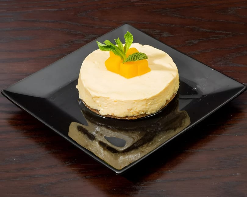 A round cheesecake with mango cubes and mint leaves on a black square plate.